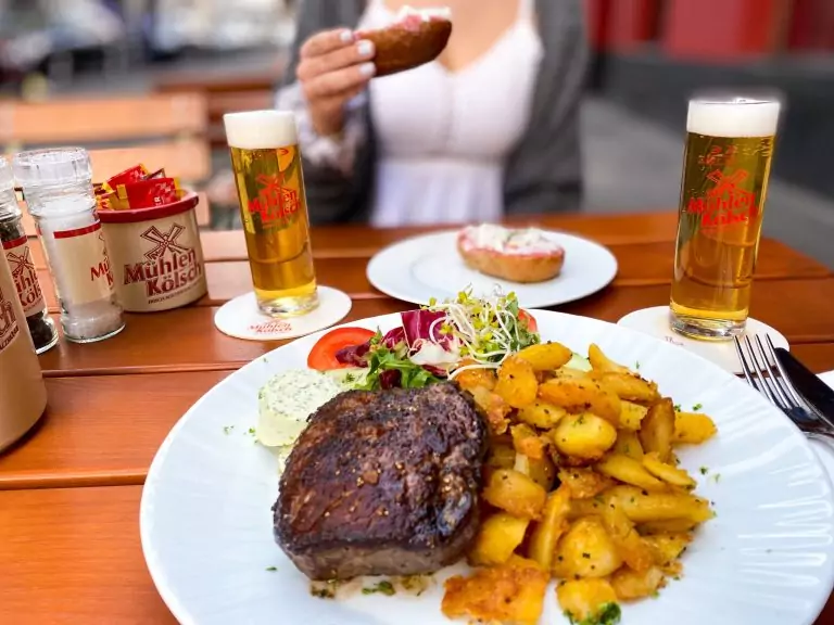 Brauhaus Köln - Brauhaus Pütz - Das Brauhaus im Belgischen Viertel. Ein Bild. Zu erkennen sind zwei Teller auf einem Tisch. Auf dem einen ist ein gebratenes Steak mit knusprigen Bratkartoffeln und Salat zu erkennen. Auf dem anderen ist ein klassischer Mett Happen zu erkennen. Dazu zwei frische Mühlen Kölsch.
